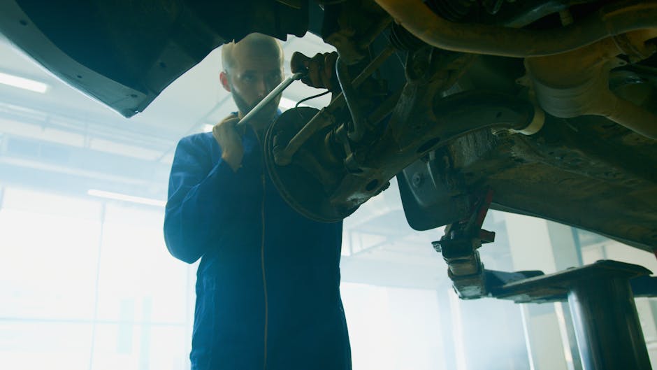 A male mechanic with a beard and short hair is working underneath a vehicle elevated on a hydraulic lift in a well-lit garage. He is wearing a blue work jumpsuit and is holding a wrench, inspecting the car's underside. The car's undercarriage displays various parts such as the exhaust system and suspension components. The garage environment features large windows allowing natural light to illuminate the workspace, and the scene reflects professional car repair and maintenance activities typical of an auto service center. This setting showcases precision in vehicle inspection, aligning with automotive repair services provided by Gardeners Kentish Town, with a focus on quality and attention to detail.